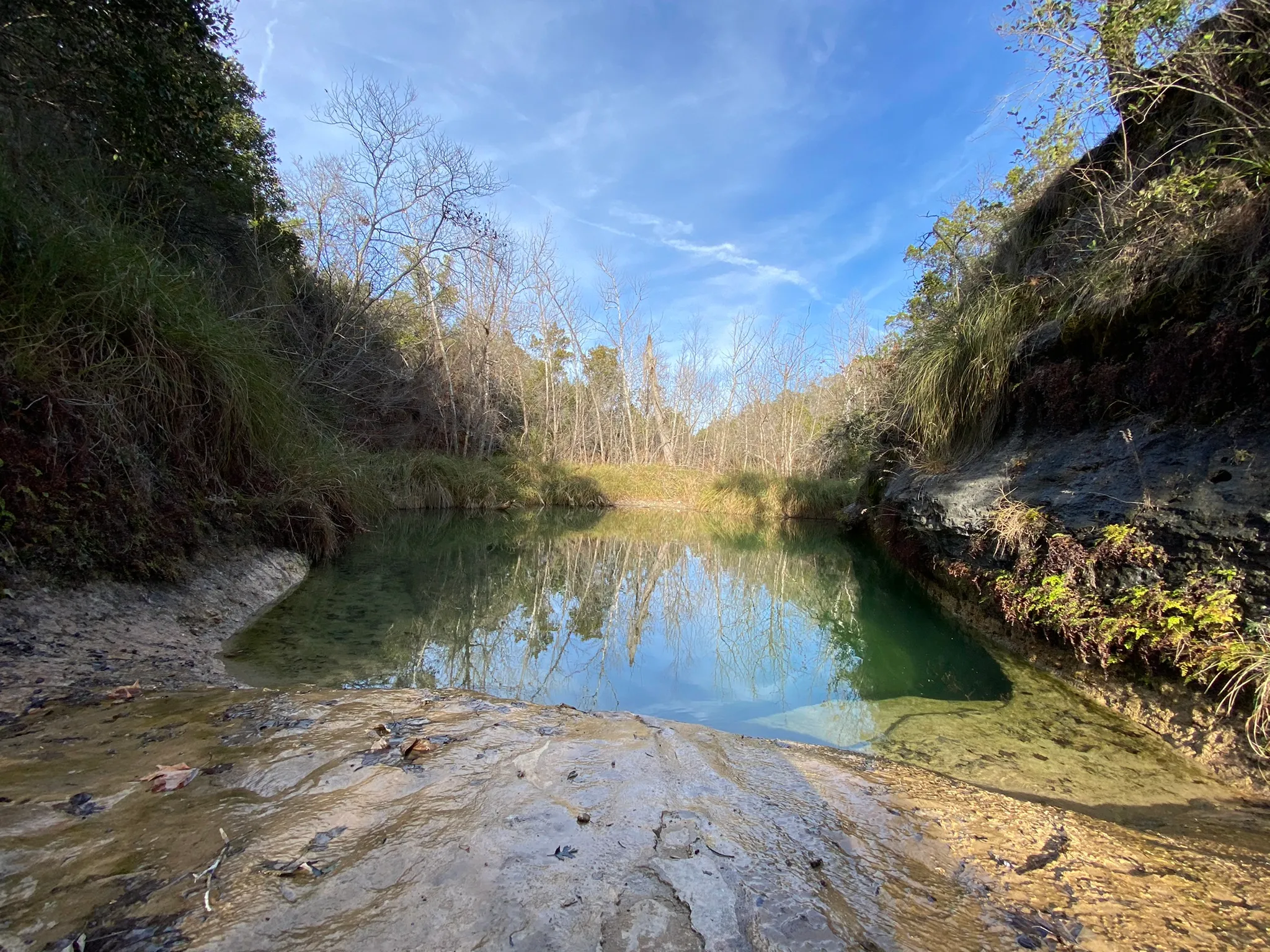 A tranquil swimming hole surrounded by rocky terrain and sparse trees under a clear blue sky.